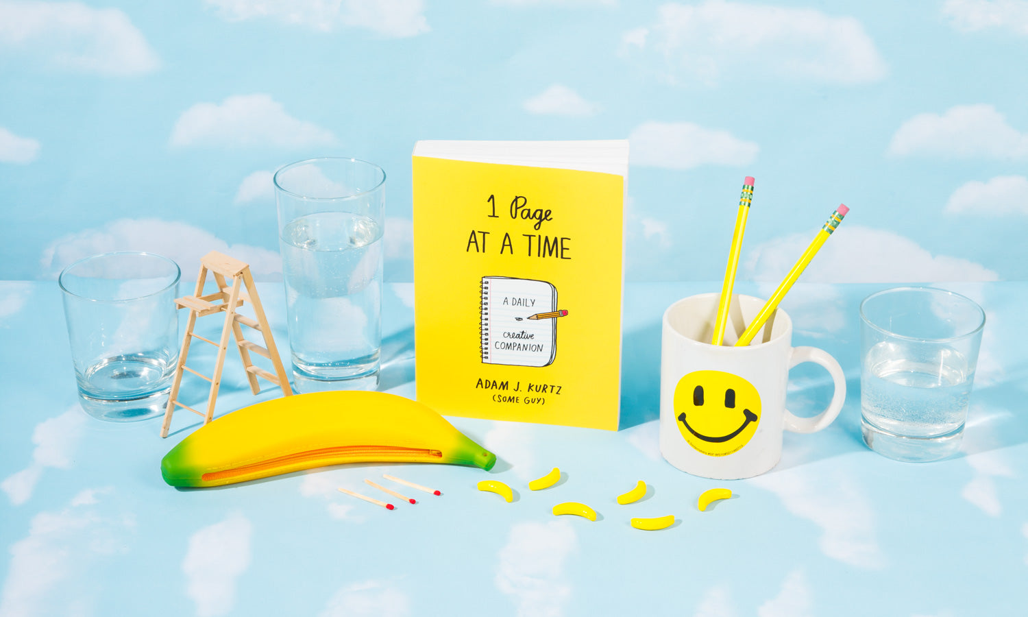 Photograph of Adam's book "1 Page at a Time" surrounded by other yellow accessories and props, against a background of painted clouds on light blue.