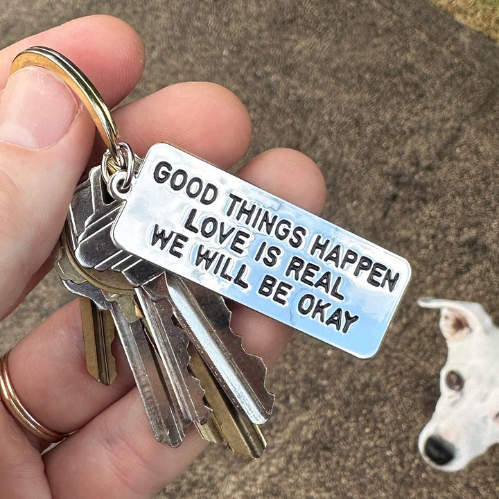 Metal keychain reads GOOD THINGS HAPPEN LOVE IS REAL WE WILL BE OKAY. Art and words by Adam JK.