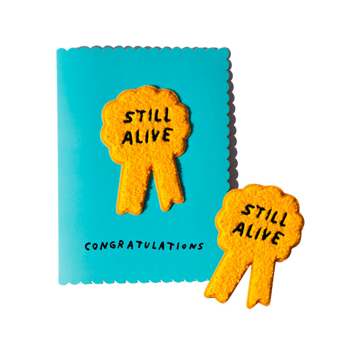 Blue greeting card with "STILL ALIVE" rosette ribbon shaped embroidery patch. By Adam JK.