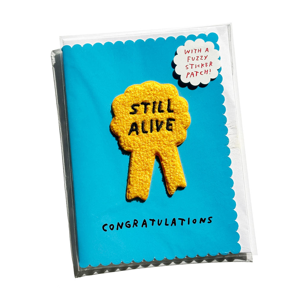 Blue greeting card with "STILL ALIVE" rosette ribbon shaped embroidery patch. By Adam JK.