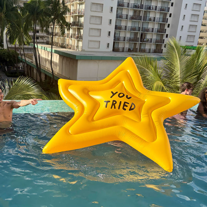 Inflatable star-shaped float with 'YOU TRIED' text in a pool setting with palm trees and buildings in the background.