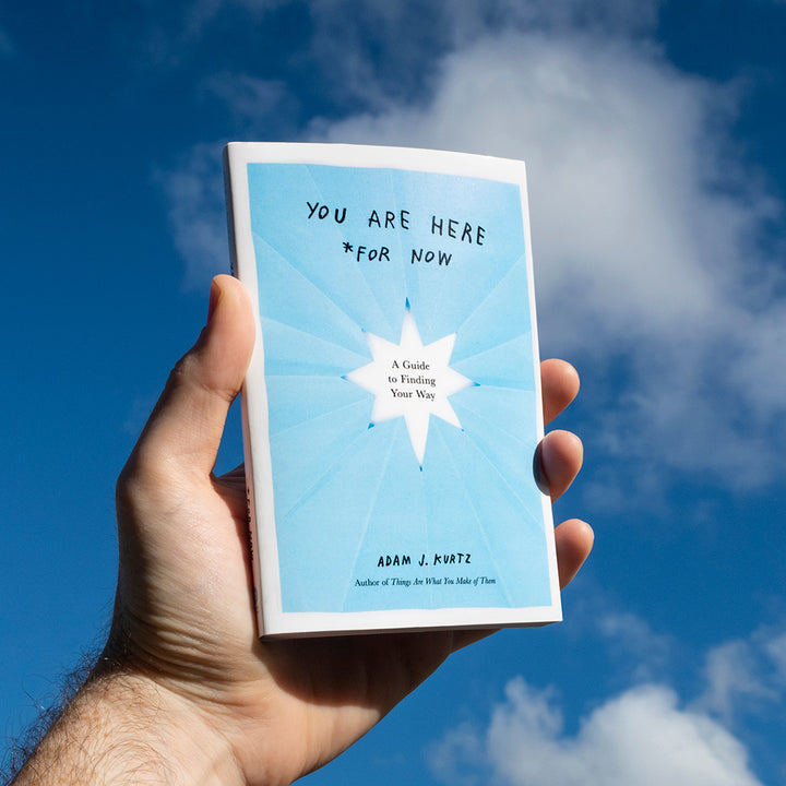 Photo of the book You Are Here (For Now) by Adam J. Kurtz held up against a blue sky with clouds.